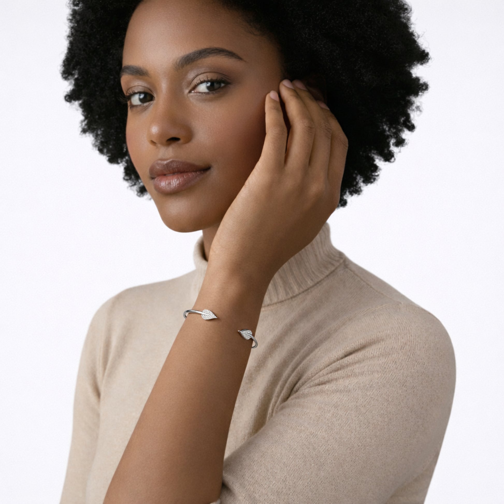 On-model portrait highlights a slim silver tone open cuff capped with pavé leaf tips, glinting against a beige turtleneck as the hand traces the jawline.