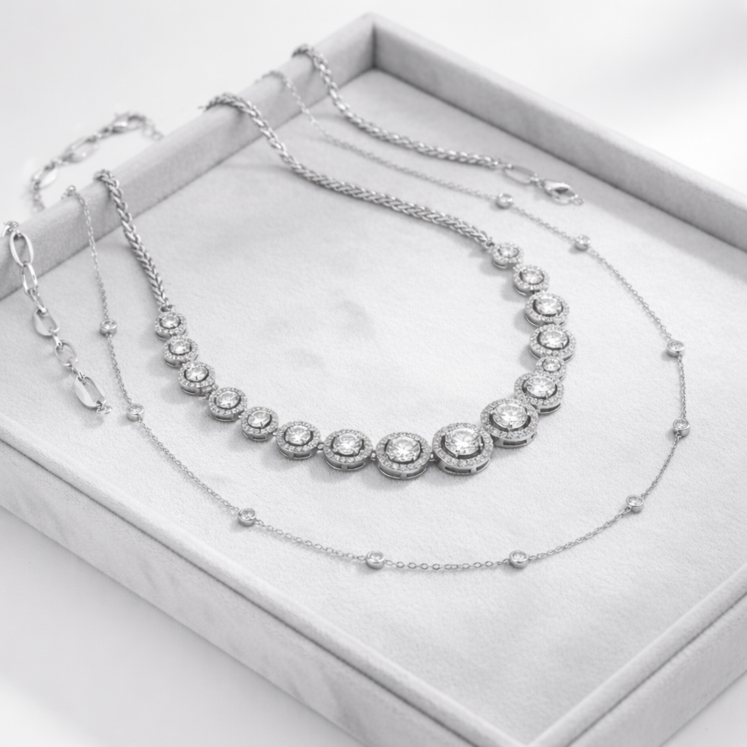 Studio view of a tennis necklace featuring round cubic zirconia stones in graduated sizes set in a silver tone finish, displayed on a soft grey jewelry tray.