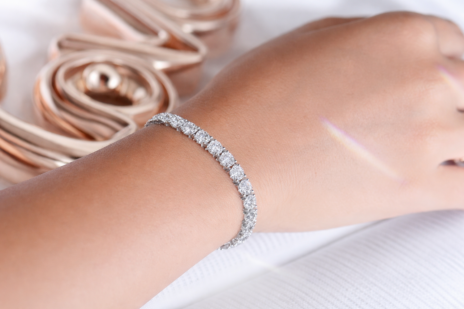 Close-up of a tennis bracelet with round cubic zirconia stones in a silver tone finish, worn on the wrist and shown against a soft fabric background.