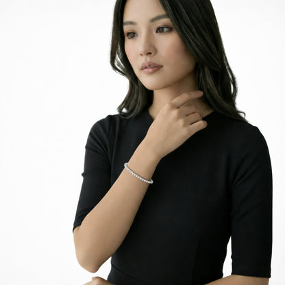 Model shot pairs a short-sleeve black dress with a slim cubic zirconia tennis bracelet, showcasing its uniform row of round stones as the hand rests near the collarbone.